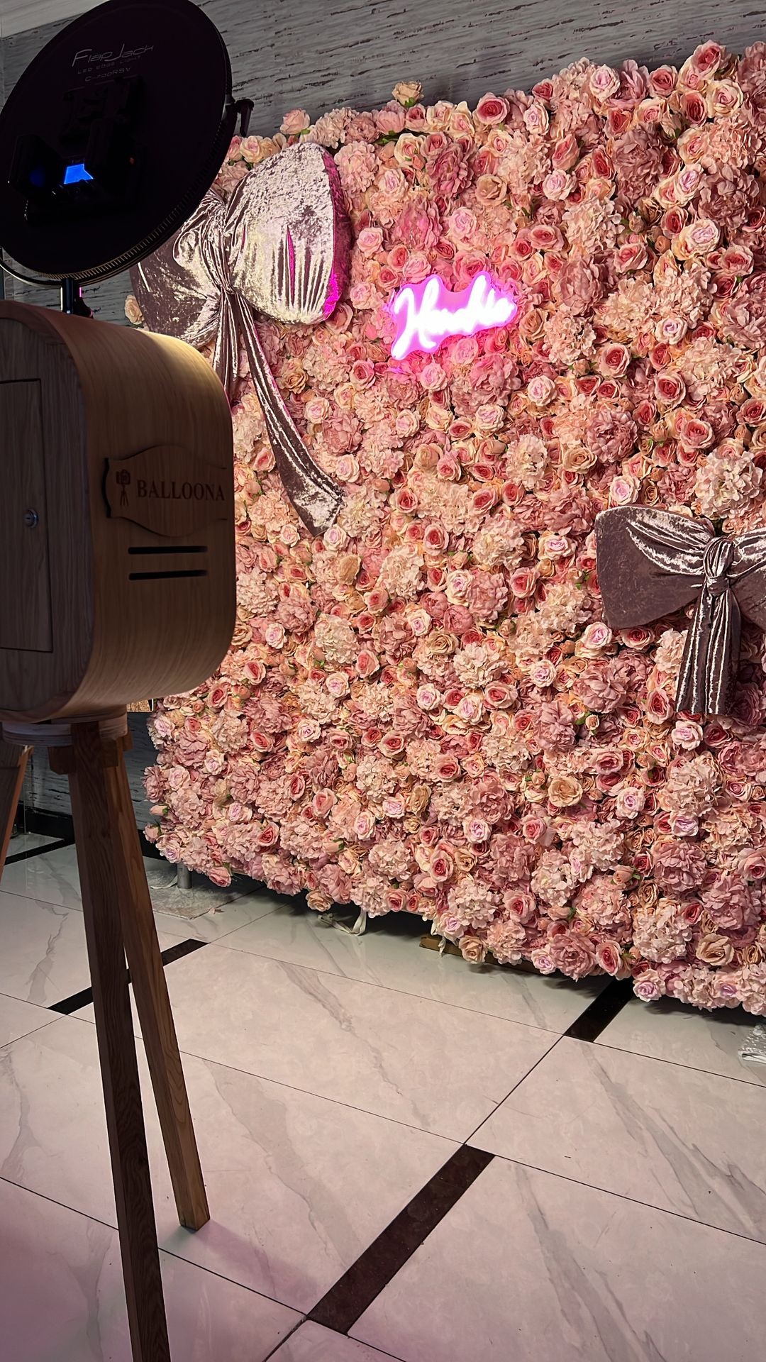 Photo booth with neon sign and floral backdrop featuring pink roses and decorative bows.